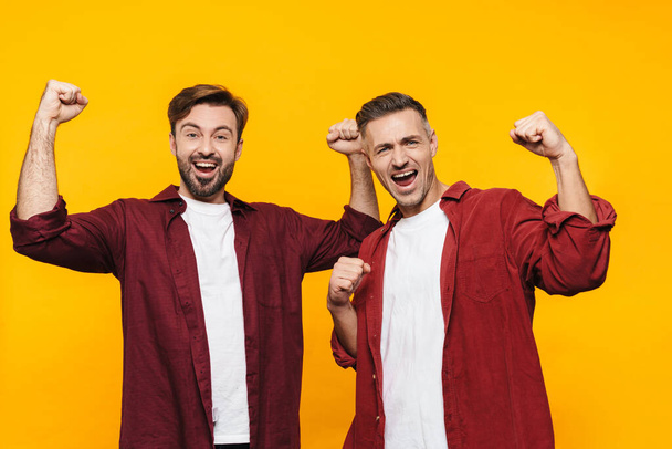Two attractive cheerful excited young men friends wearing casual clothes standing isolated over yellow background, flexing biceps - Fotó, kép