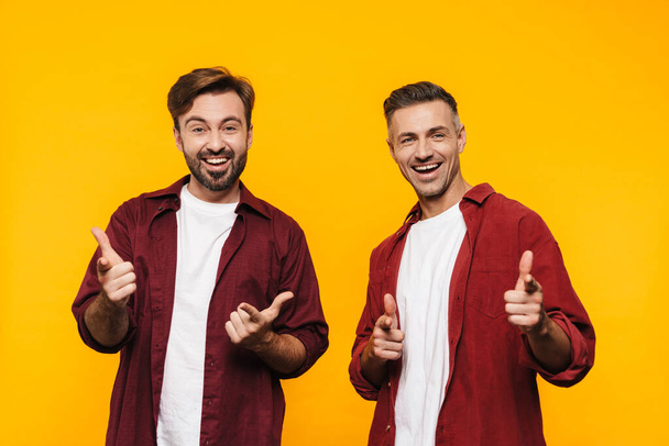 Two attractive cheerful excited young men friends wearing casual clothes standing isolated over yellow background, pointing fingers at camera - 写真・画像
