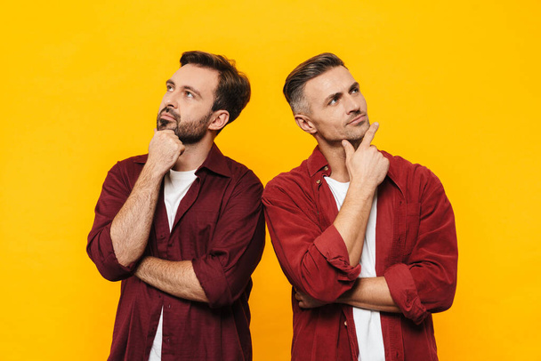 Image of two handsome men 30s in red shirts thinking and looking upwards at copyspace isolated over yellow background - Foto, Imagem