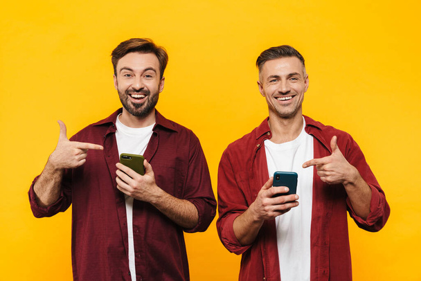 Image of two handsome men 30s in red shirts smiling and pointing fingers at cellphones isolated over yellow background - Zdjęcie, obraz