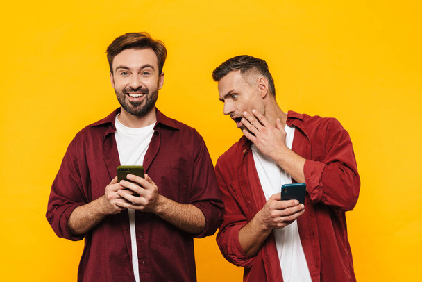 Image of handsome men 30s in red shirts holding and peeking at cellphone isolated over yellow background - Zdjęcie, obraz