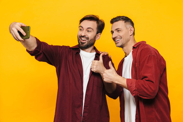 Two attractive cheerful excited young men friends wearing casual clothes standing isolated over yellow background, taking a selfie, thumbs up - Fotografie, Obrázek