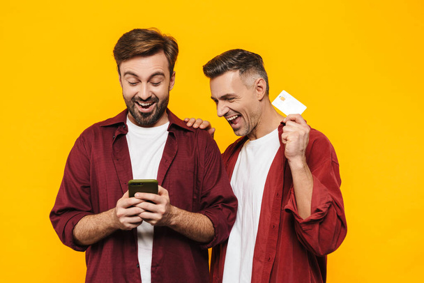 Two attractive cheerful excited young men friends wearing casual clothes standing isolated over yellow background, shopping online with mobile phone and credit card - Fotoğraf, Görsel