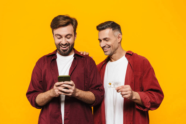 Two attractive cheerful excited young men friends wearing casual clothes standing isolated over yellow background, shopping online with mobile phone and credit card - Foto, Bild