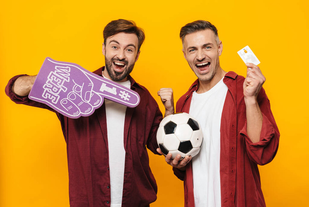 Two attractive cheerful excited young men friends soccer fans wearing casual clothes standing isolated over yellow background, showing credit card - Fotografie, Obrázek