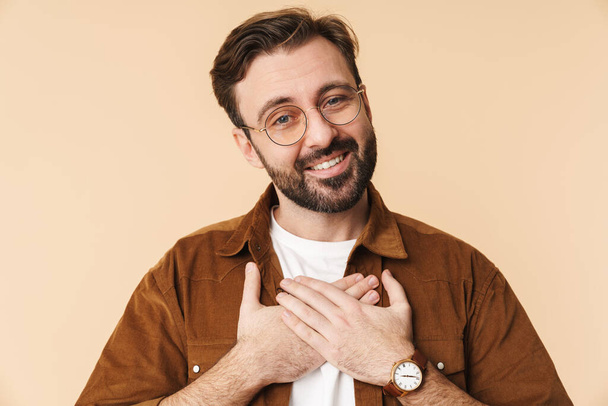 Portrait of a cheerful young arttractive bearded man wearing casual clothes standing isolated over beige background, holding hands at his chest - Фото, зображення