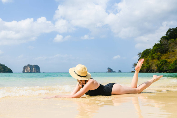 Şapkalı bir kız, güzel kayalara ve denize bakıyor, siyah mayo giyiyor, bacak kaldırıyor ve Tayland 'da tropikal tatilin tadını çıkarıyor. - Fotoğraf, Görsel