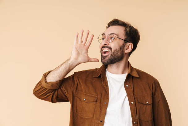 Portrait of a cheerful young arttractive bearded man wearing casual clothes standing isolated over beige background, yelling loud - Foto, Bild