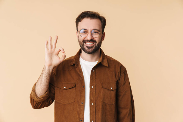 Portrait of a cheerful young arttractive bearded man wearing casual clothes standing isolated over beige background, showing ok gesture - Foto, Bild