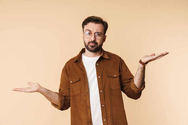Portrait of a confused young arttractive bearded man wearing casual clothes standing isolated over beige background, shrugging shoulders - Foto, Bild