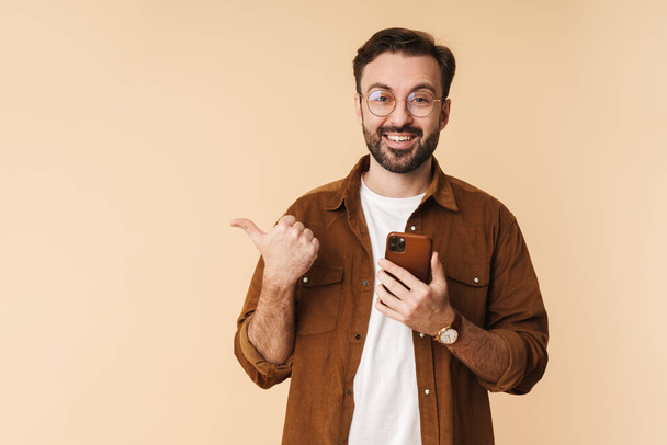 Portrait of a cheerful young arttractive bearded man wearing casual clothes standing isolated over beige background, using mobile phone, pointing finger away at copy space - Foto, Imagen