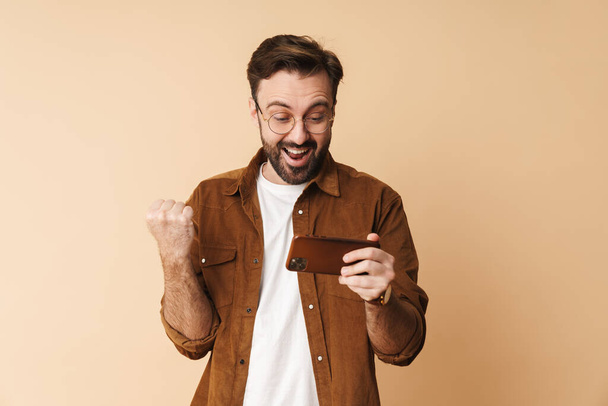 Portrait of a cheerful young arttractive bearded man wearing casual clothes standing isolated over beige background, using mobile phone, celebrating success - Photo, image