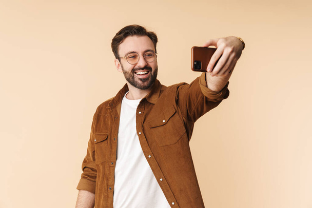 Retrato de un joven y alegre hombre barbudo arttractivo con ropa casual de pie aislado sobre fondo beige, tomando una selfie
 - Foto, Imagen