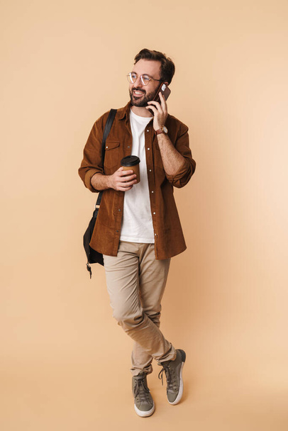 Full length portrait of a cheerful young arttractive bearded man wearing casual clothes standing isolated over beige background, talking on mobile phone, holding takeaway coffee cup - 写真・画像
