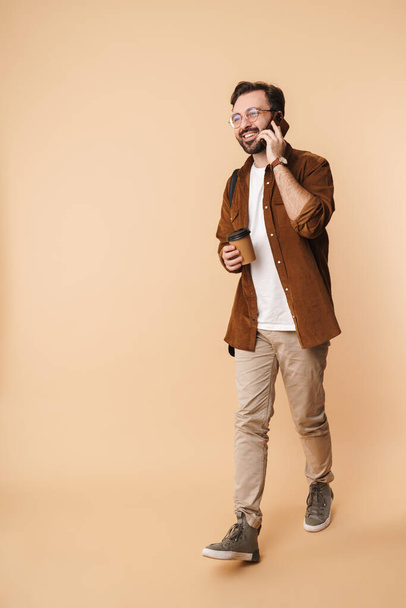 Full length portrait of a cheerful young arttractive bearded man wearing casual clothes walking isolated over beige background, talking on mobile phone, holding takeaway coffee cup - Fotó, kép