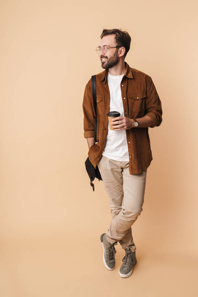 Full length portrait of a cheerful young arttractive bearded man wearing casual clothes standing isolated over beige background, holding takeaway coffee cup - Фото, изображение