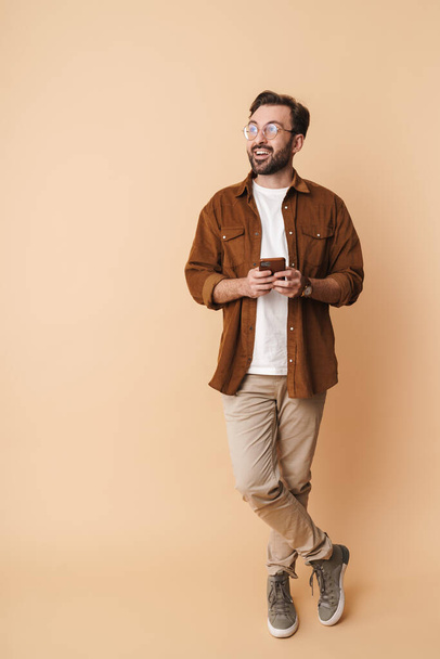 Full length portrait of a cheerful young arttractive bearded man wearing casual clothes standing isolated over beige background, holding mobile phone, looking away - Foto, immagini