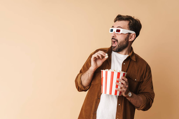 Portrait of a cheerful young arttractive bearded man wearing casual clothes standing isolated over beige background, watching a movie in 3d glasses, eating popcorn, looking away - Zdjęcie, obraz