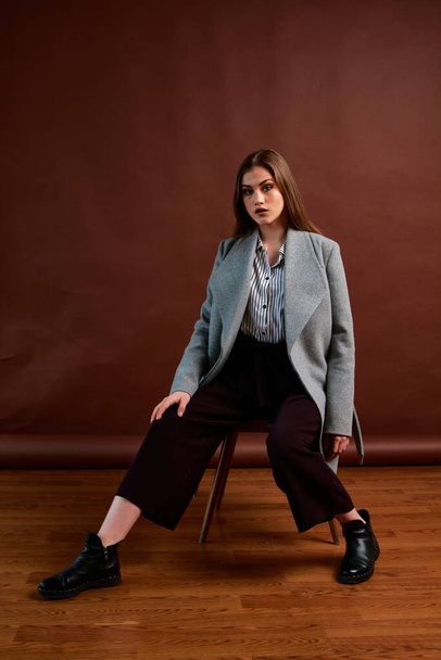 Independent young woman posing in the photostudio. Gorgeous young female in classic suit and grey coat. Brown background. Vintage chair style. Woman dressed in Man clothes. Fashionable clothes. - Foto, imagen