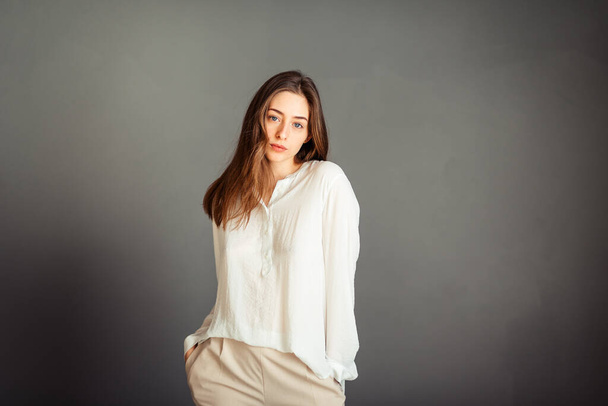 Portret van een jong meisje in een wit shirt, recht kijkend, beide handen in de zak op een grijze achtergrond. Geen retoucheren. Zonder make-up. - Foto, afbeelding
