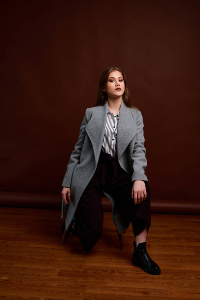 Independent young woman posing in the photostudio. Gorgeous young female in classic suit and grey coat. Brown background. Vintage chair style. Woman dressed in Man clothes. Fashionable clothes. - Фото, зображення