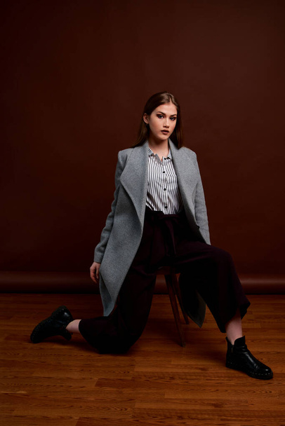 Independent young woman posing in the photostudio. Gorgeous young female in classic suit and grey coat. Brown background. Vintage chair style. Woman dressed in Man clothes. Fashionable clothes. - Foto, Bild