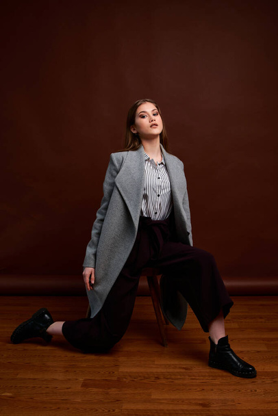 Independent young woman posing in the photostudio. Gorgeous young female in classic suit and grey coat. Brown background. Vintage chair style. Woman dressed in Man clothes. Fashionable clothes. - Fotó, kép