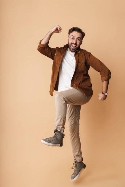 Full length portrait of a cheerful young arttractive bearded man wearing casual clothes jumping isolated over beige background, celebrating success - 写真・画像