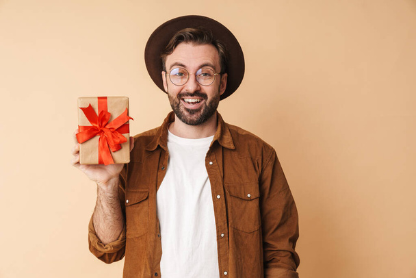 Image of a happy excited young unshaved man in hat isolated over beige wall background holding present box. - 写真・画像