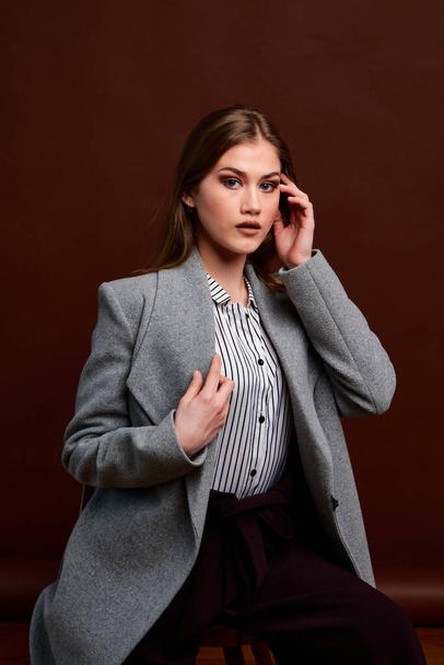 Independent young woman posing in the photostudio. Gorgeous young female in classic suit and grey coat. Brown background. Vintage chair style. Woman dressed in Man clothes. Fashionable clothes. - Photo, Image