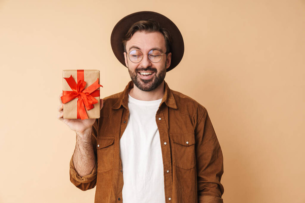 Image of a happy optimistic young unshaved man in hat isolated over beige wall background holding present box. - Фото, зображення