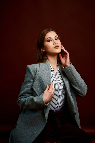 Independent young woman posing in the photostudio. Gorgeous young female in classic suit and grey coat. Brown background. Vintage chair style. Woman dressed in Man clothes. Fashionable clothes. - Fotografie, Obrázek