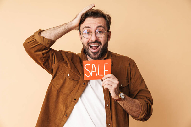 Image of a happy excited surprised young unshaved man isolated over beige wall background holding sale blank. - Zdjęcie, obraz