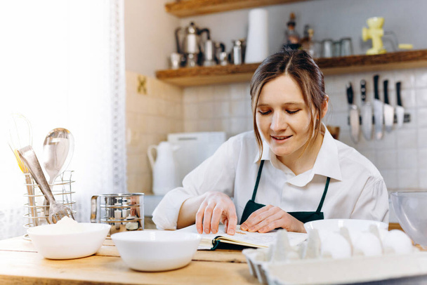 Immagine ravvicinata di donna seduta al tavolo da cucina in legno che cerca di ricettario e cerca di scegliere cosa cucinare. Cucinare a casa concetto, stile di vita. Dieta chetogenica e menu
 - Foto, immagini