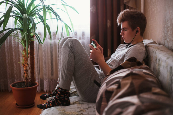 A young guy, a teenager sit on the sofa at home in headphone and writes sms on a social network or listen music on a smartphone. - Foto, Imagen