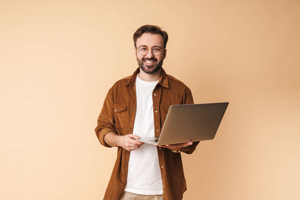 Image of a happy cheery optimistic young unshaved man isolated over beige wall background using laptop computer. - Foto, afbeelding