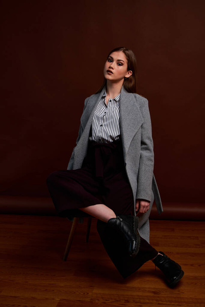Independent young woman posing in the photostudio. Gorgeous young female in classic suit and grey coat. Brown background. Vintage chair style. Woman dressed in Man clothes. Fashionable clothes. - Fotografie, Obrázek