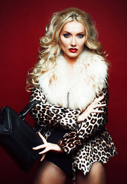 Beautiful fashionable woman with blond hair, dressed in a leopard jacket, holds a bag and poses on a red background. - Fotoğraf, Görsel