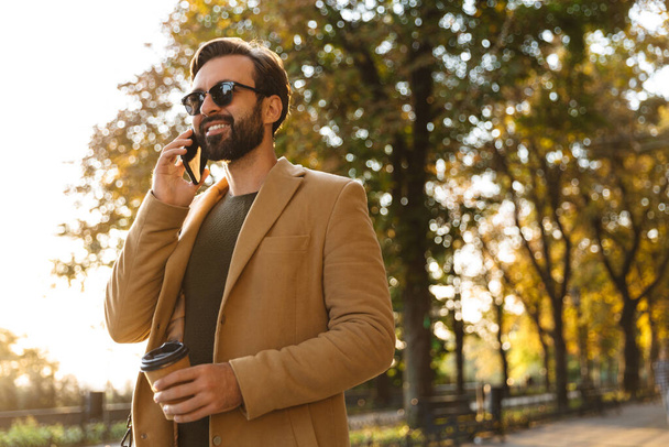Image of handsome adult man in jacket smiling and talking on smartphone while walking in park - Fotoğraf, Görsel