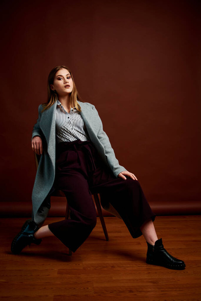 Independent young woman posing in the photostudio. Gorgeous young female in classic suit and grey coat. Brown background. Vintage chair style. Woman dressed in Man clothes. Fashionable clothes. - Foto, Bild