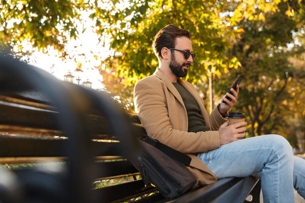 Image of handsome adult man in jacket using cellphone while sitting on bench in park - Photo, Image
