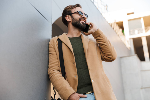Image of handsome caucasian bearded adult man in sunglasses talking on smartphone and standing by building outdoors - Foto, afbeelding