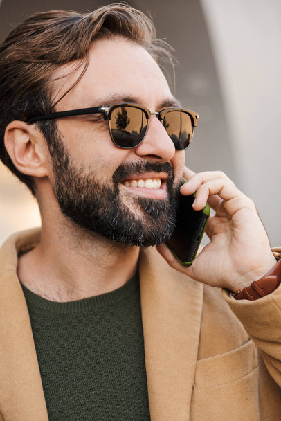 Kép jóképű fehér szakállas felnőtt férfi napszemüvegben beszél okostelefonon, és álló épület szabadban - Fotó, kép