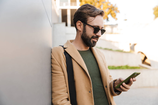 Image of handsome caucasian bearded adult man in sunglasses holding smartphone and standing by building outdoors - Fotografie, Obrázek