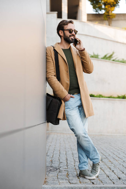 Image of handsome caucasian bearded adult man in sunglasses talking on smartphone and standing by building outdoors - Fotó, kép