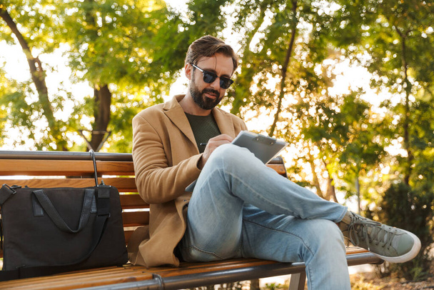 Image of handsome adult man in jacket woking and holding clipboard while sitting on bench in park - Photo, Image