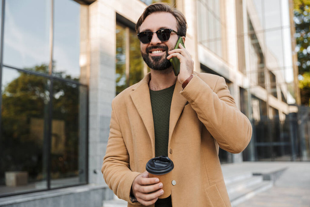 Image of handsome bearded adult man in sunglasses talking on smartphone while walking at city street with coffee cup - Fotó, kép