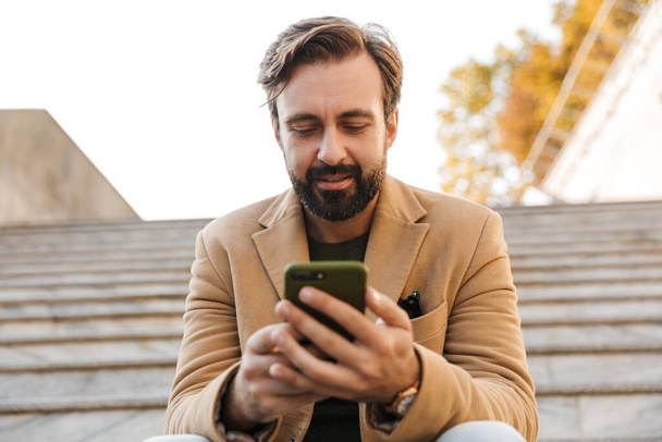 Image of handsome adult man in jacket holding smartphone while sitting on stairs outdoors - Foto, Imagen