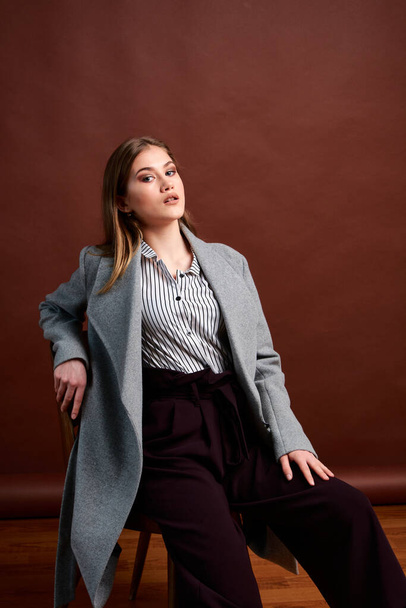 Independent young woman posing in the photostudio. Gorgeous young female in classic suit and grey coat. Brown background. Vintage chair style. Woman dressed in Man clothes. Fashionable clothes. - Фото, изображение