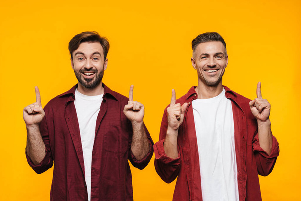 Image of two handsome men 30s in red shirts smiling and pointing fingers upward at copyspace isolated over yellow background - Photo, Image